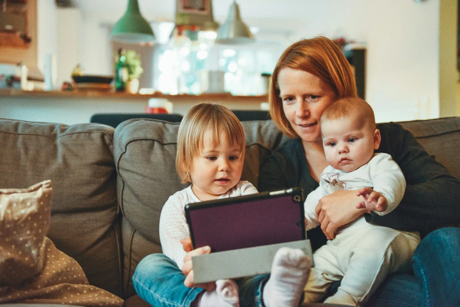 Mutter mit zwei kleinen Kindern auf dem Sofa, die gemeinsam auf ein Tablet schauen